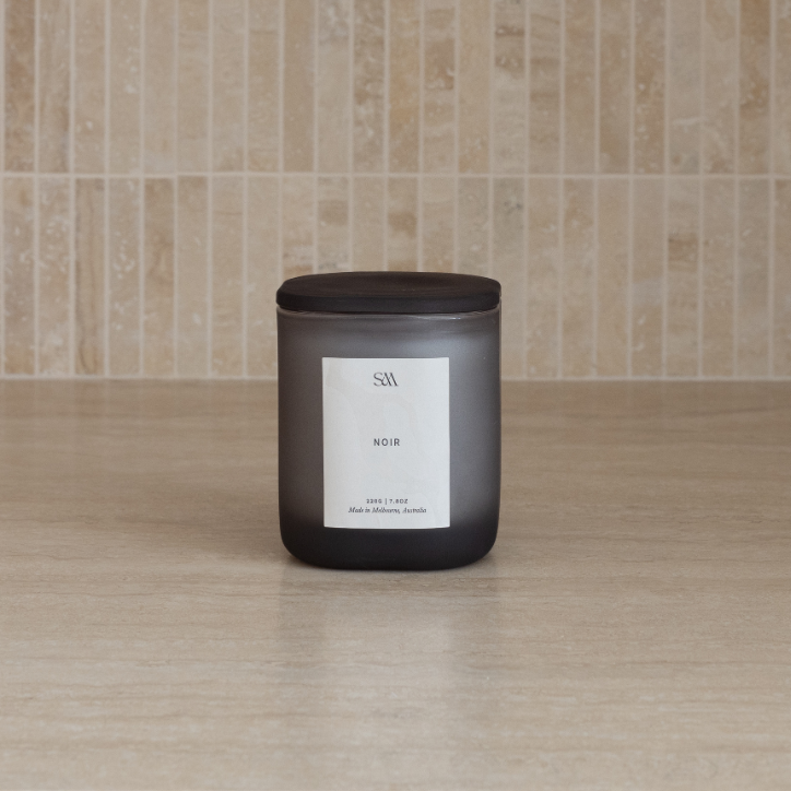 A dark frosted glass candle jar with a black lid and a minimalist white label, set on a light wood surface against a beige tiled wall.