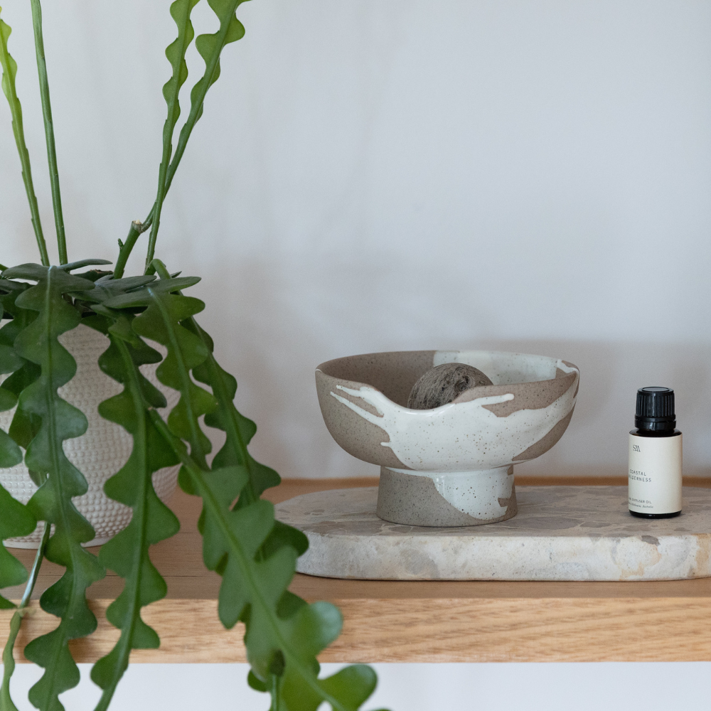 Ceramic bowl with a stone on a wooden shelf next to a plant and a bottle of essential oil.