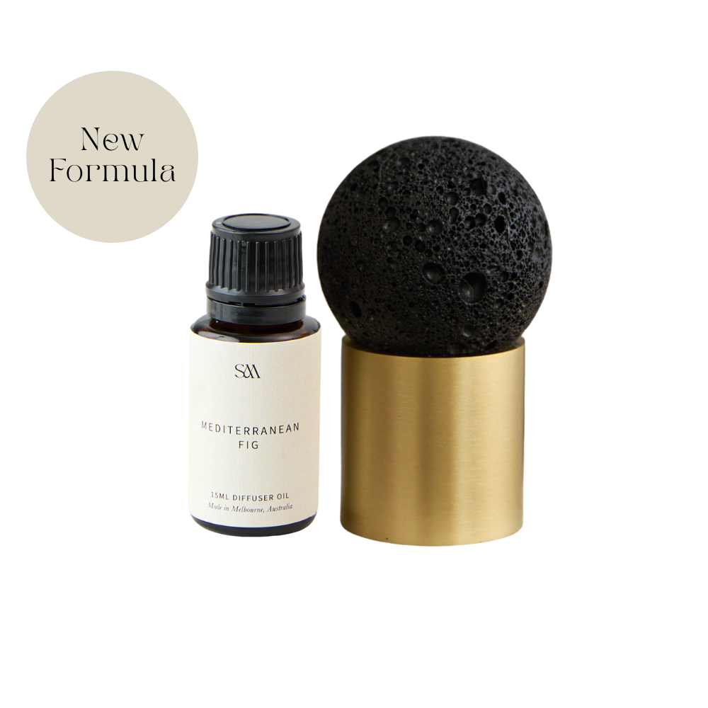 A small fragrance oil bottle with a cream label and black cap stands beside a black porous stone sphere resting on a brushed brass cylindrical base. A beige circle in the upper left says 'New Formula.