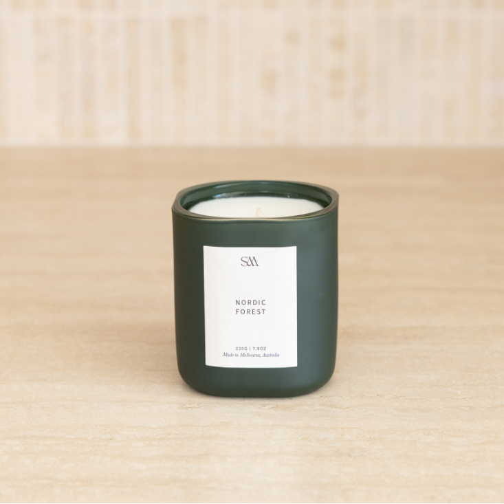 A dark green glass jar candle with a white label reading 'SM Nordic Forest' sits on a light wood surface against a matching light wood background.