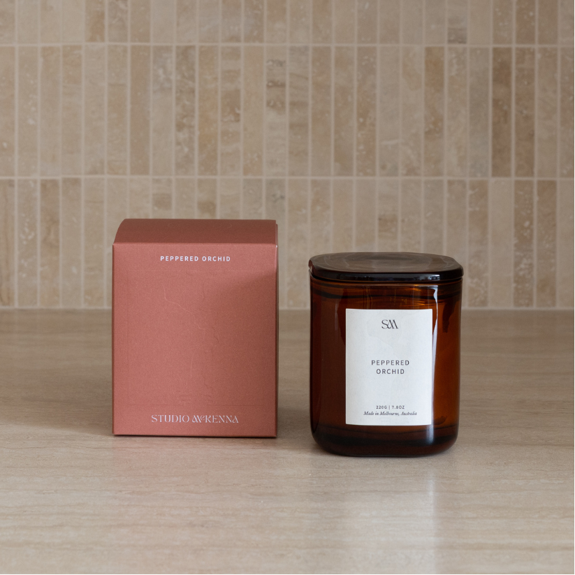 An amber glass jar candle with a lid and a white label sits next to its dusty rose box on a light wood surface against a beige tiled wall. The labels read 'STUDIO MCKENNA' and 'PEPPERED ORCHID'.