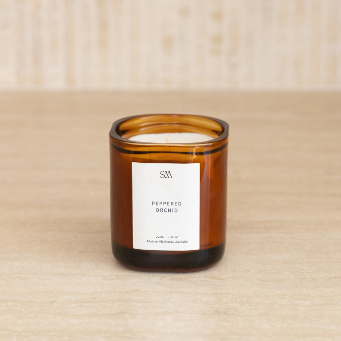 An amber glass jar candle with a white label reading 'PEPPERED ORCHID' sits on a light wood surface against a beige tiled background.