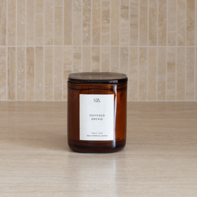 An amber glass jar candle with a white label reading 'PEPPERED ORCHID' and a glass lid, placed on a light beige countertop against a beige tiled backsplash.