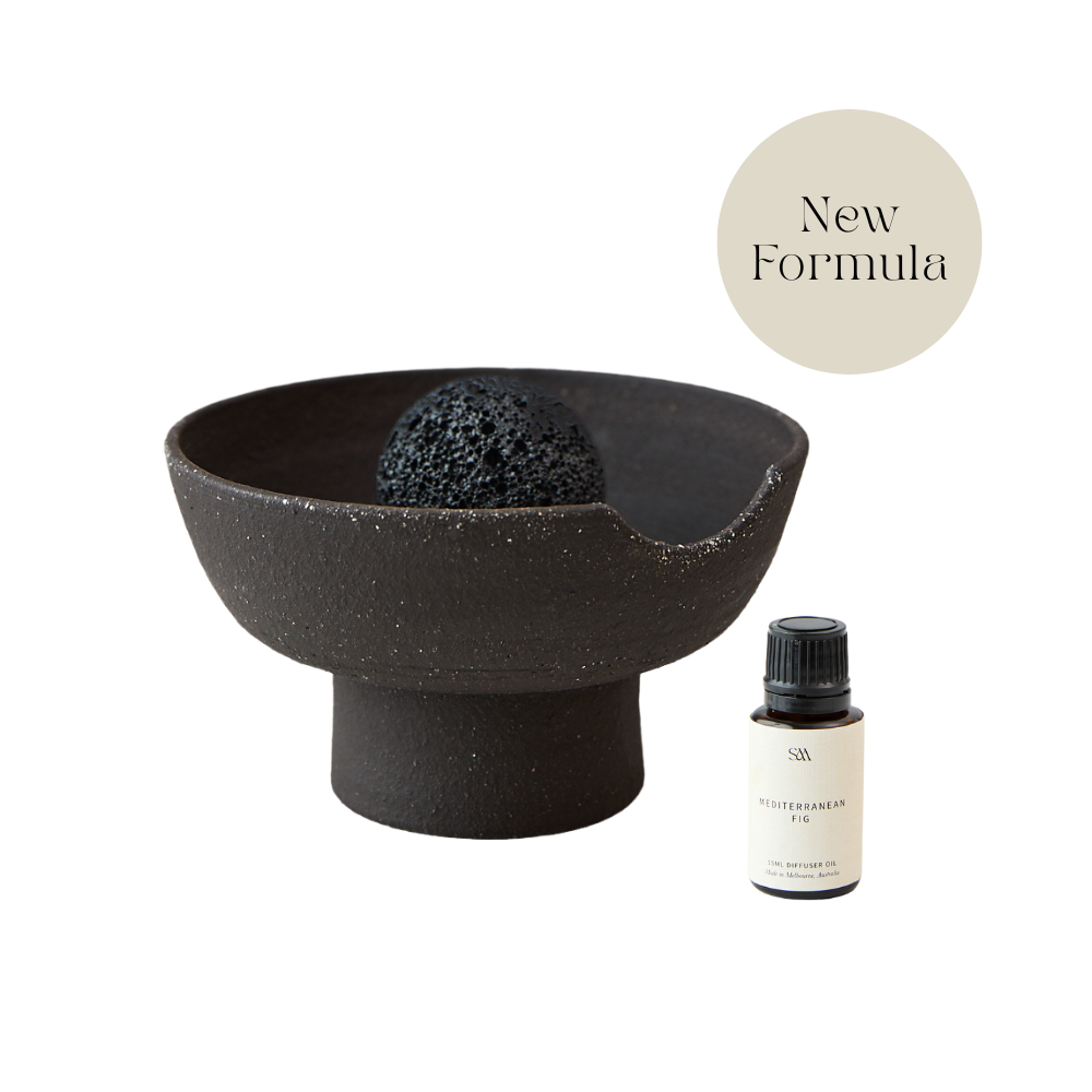 A dark charcoal-colored ceramic bowl with a textured, speckled finish, containing a black volcanic stone. Next to it is a small bottle of 15ml fragrance oil, and a beige circular badge in the top right corner reads 'New Formula'.